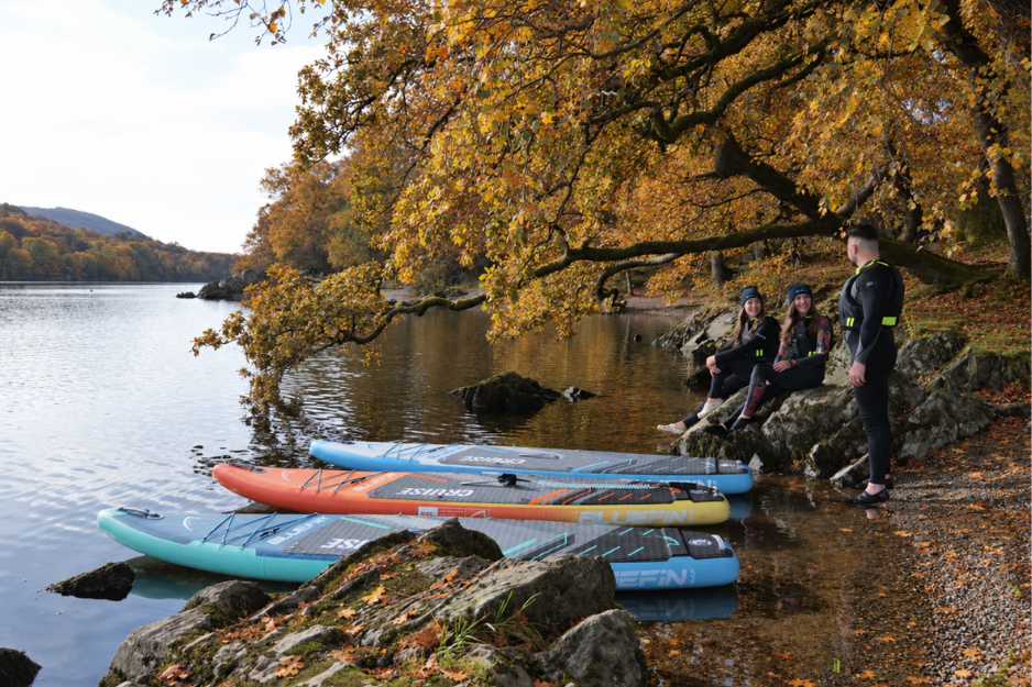 three people sat by the edge of a lake with their boards pointing towards the water on the shore
