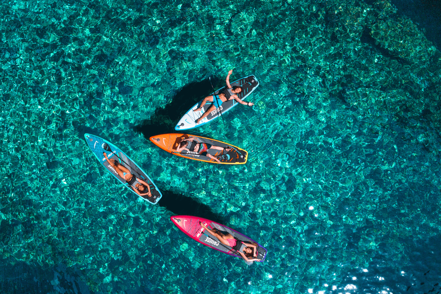 Four paddleboarders on colorful boards in clear turquoise water, enjoying a sunny day.