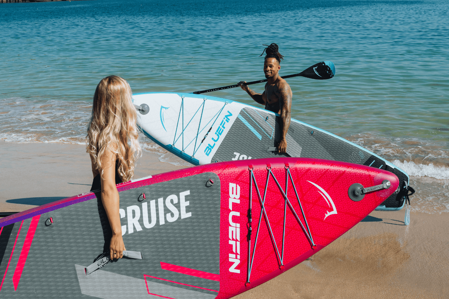 Two people on a beach holding inflatable paddleboards, one pink and one blue, with water in the background.