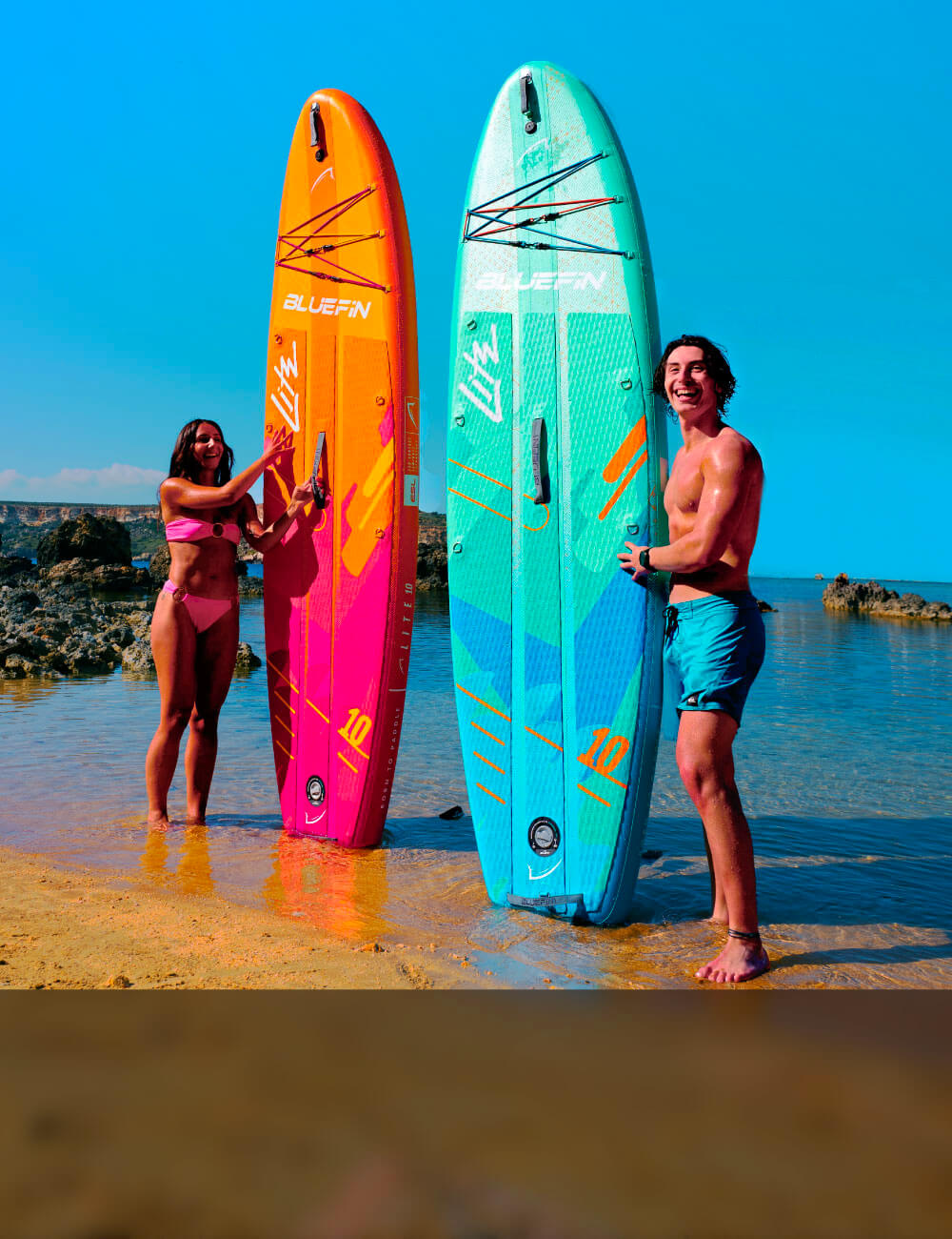 Two inflatable paddleboards in vibrant colors, one orange and one teal, on a sandy beach with two people.