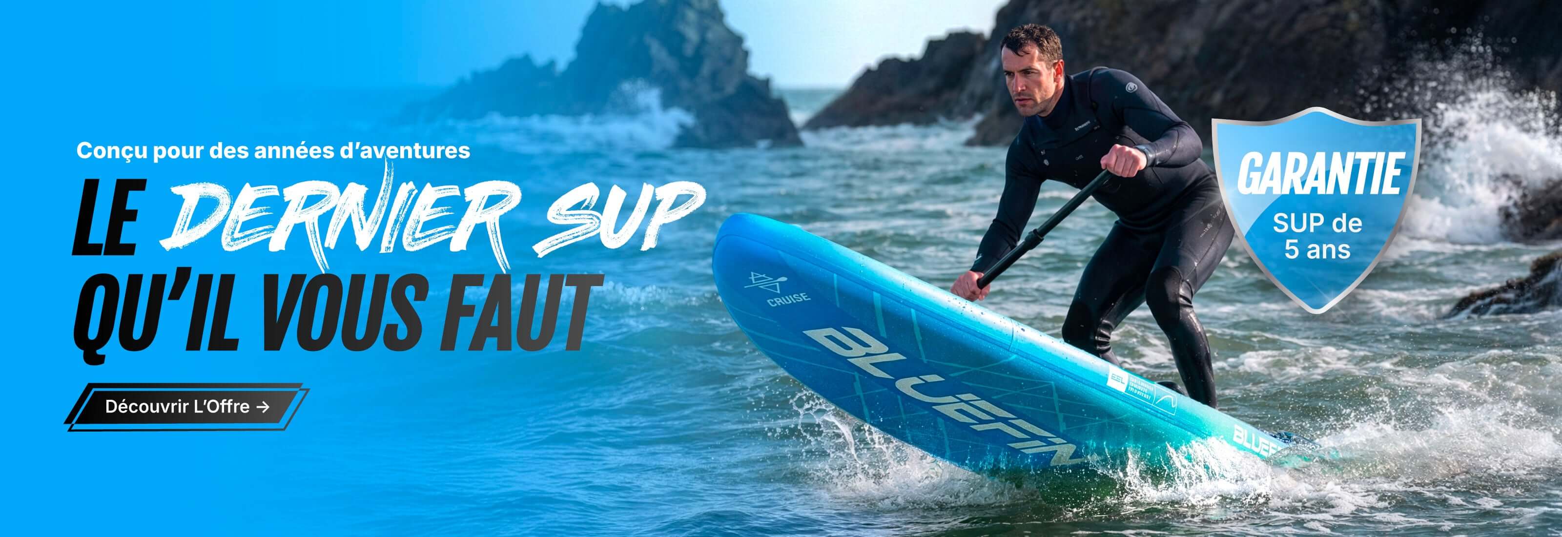 Man in wetsuit paddling a blue inflatable SUP on water with rocky shore in background