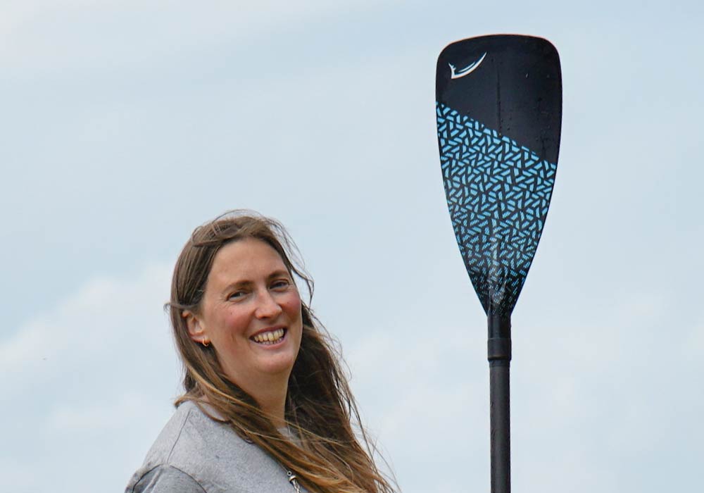 Smiling woman holding a black paddle with blue geometric design against a cloudy sky.