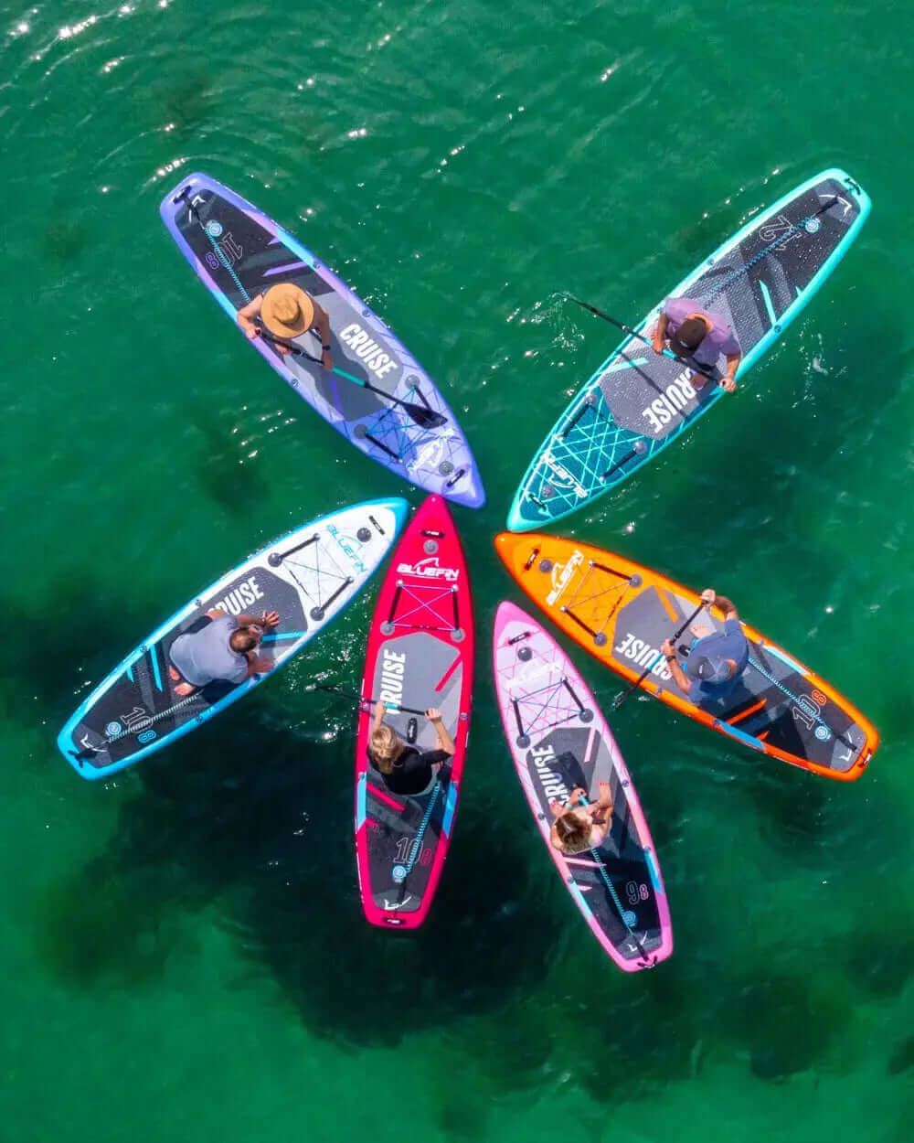 Six colorful paddleboards arranged in a circle on clear water, with paddlers using them.