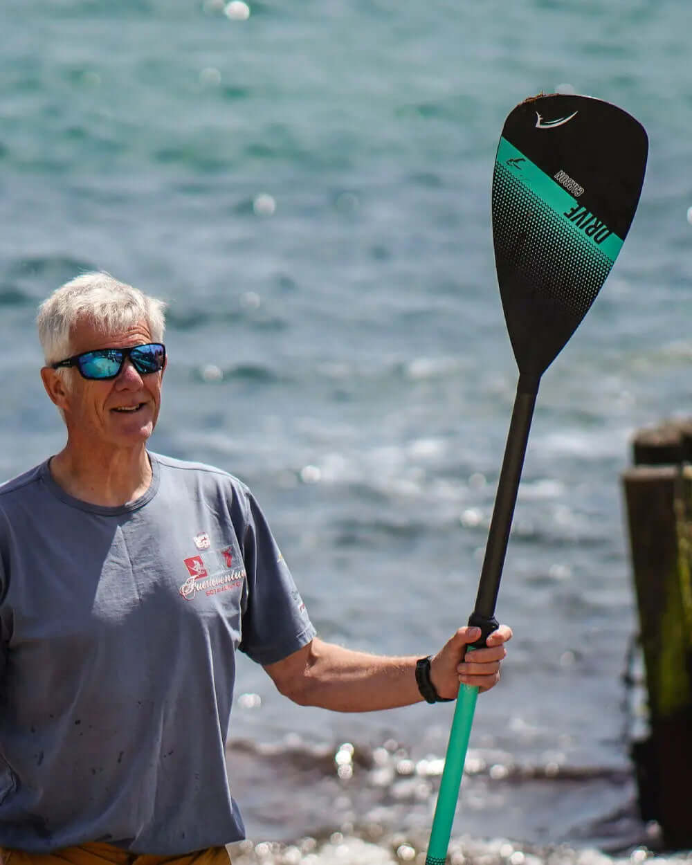 Man holding a black and teal paddle near the water, wearing sunglasses and a gray shirt