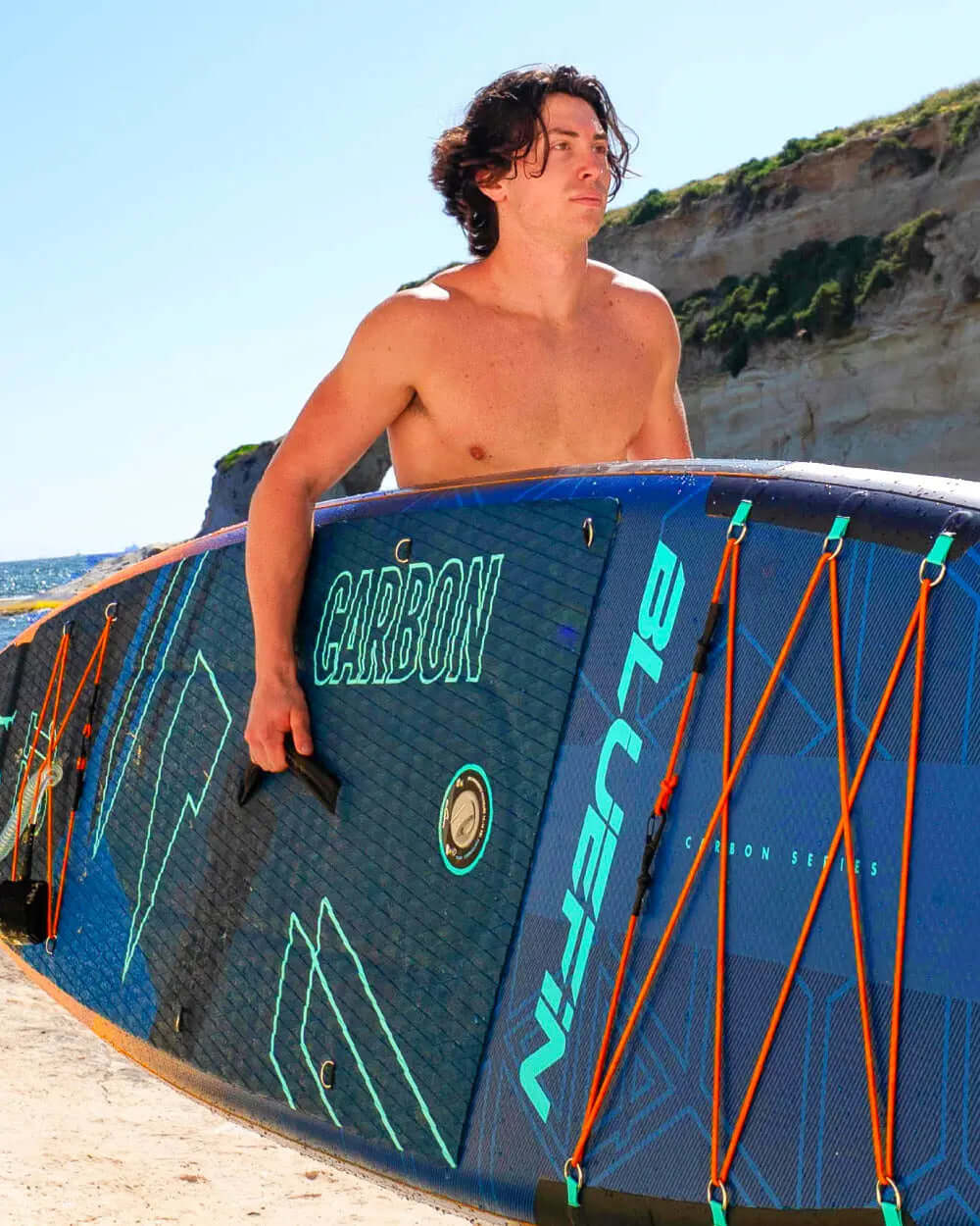 Man carrying a Bluefin SUP Carbon Series paddleboard on a beach with cliffs in the background