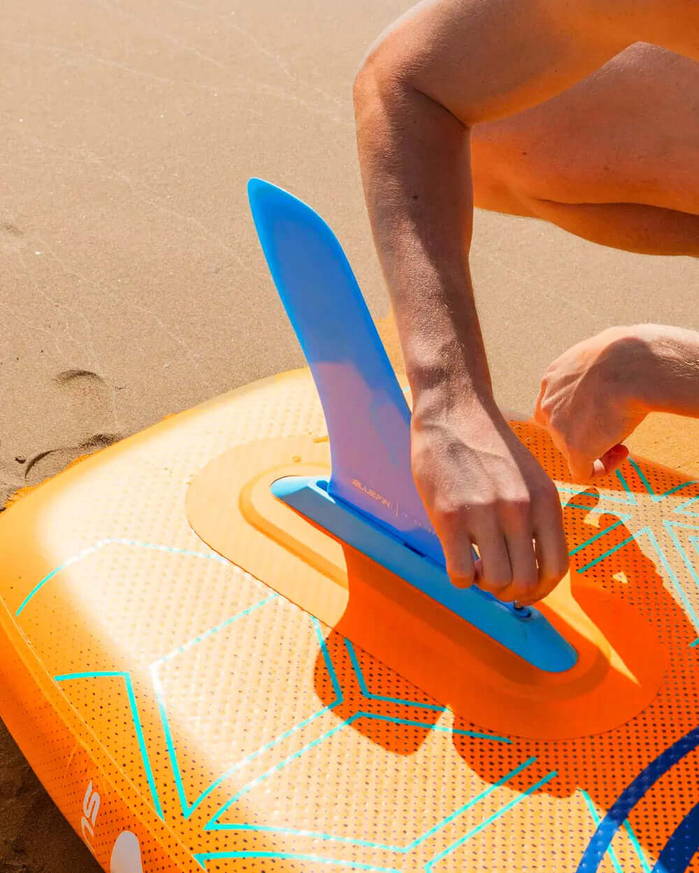 Inflatable paddleboard with orange design, blue fin being attached on sandy beach
