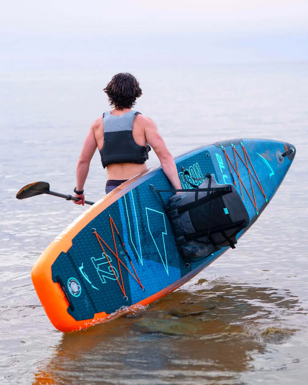 Person carrying a blue inflatable paddleboard with orange accents and a backpack, walking in shallow water