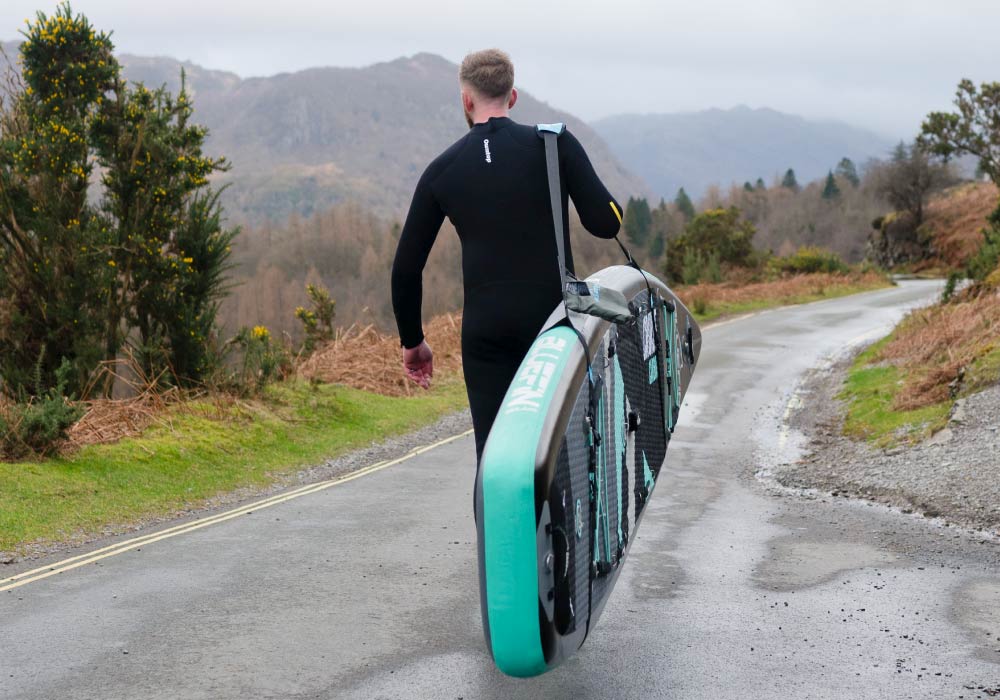 Person in wetsuit carrying an inflatable paddleboard along a winding road in a scenic landscape.