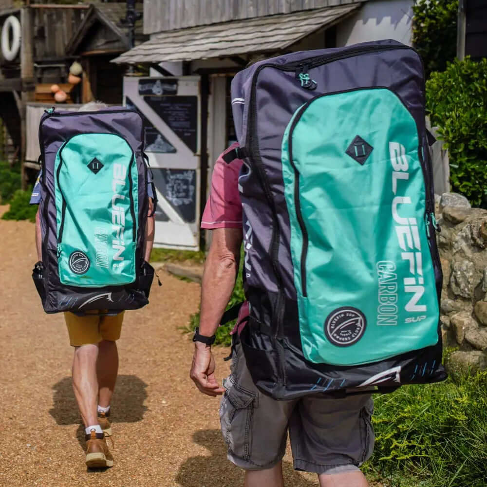 Two people walking with large Bluefin Carbon SUP backpacks on a gravel path near buildings.