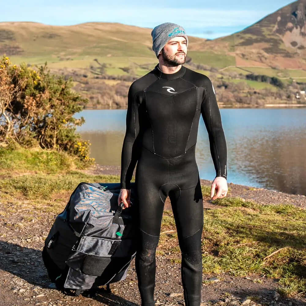 Man in black wetsuit carrying a wheeled bag by a lake with hills in the background