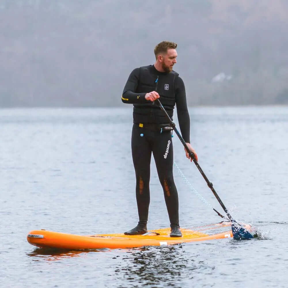 Homme en combinaison de plongée pagayant sur un paddleboard orange sur une eau calme.