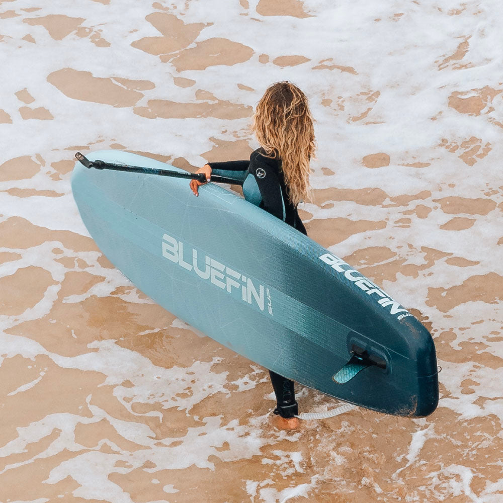 Person in wetsuit carrying a Bluefin paddleboard on a sandy beach with waves.