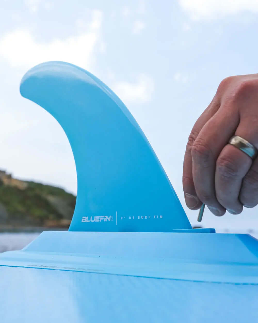 Hand attaching a blue 9" surf fin to a paddleboard with a textured surface.
