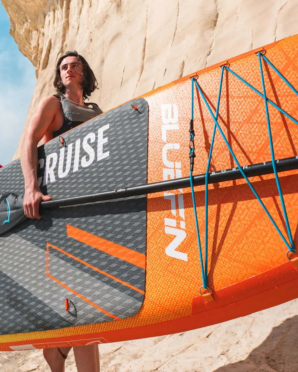 Person carrying an inflatable paddleboard with orange and gray design near a rocky beach