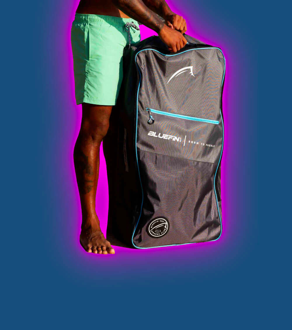 Person holding a large Bluefin bag with logo, wearing green shorts and standing barefoot on a colorful background.