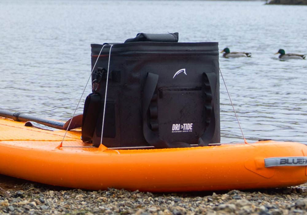 Black waterproof cooler bag on an orange paddleboard by the water with ducks in the background
