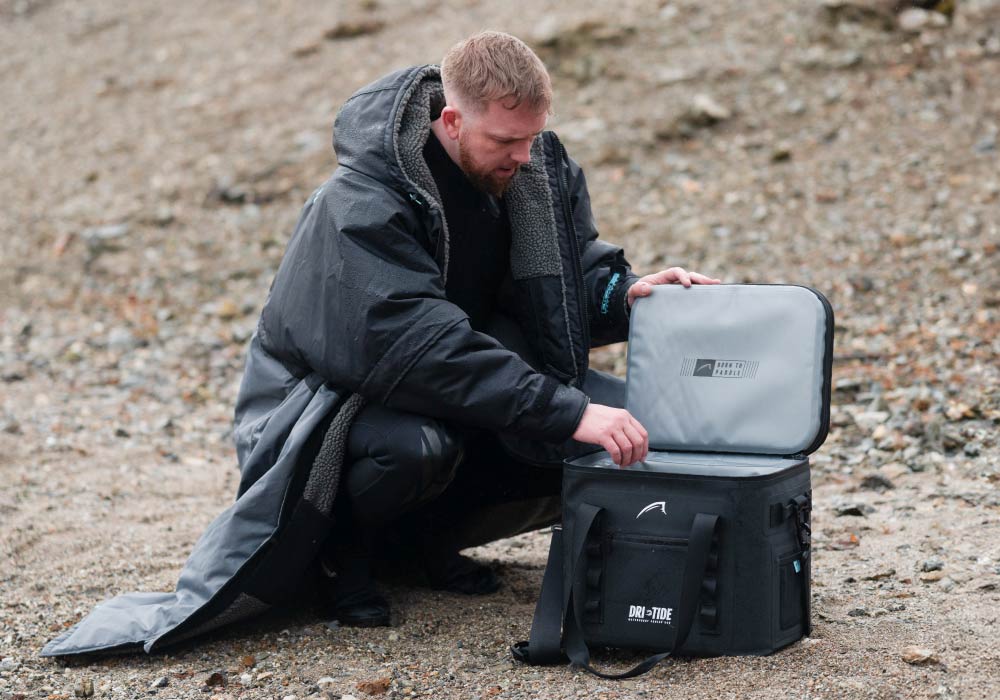Person in a black coat opening a gray cooler bag on a rocky surface outdoors