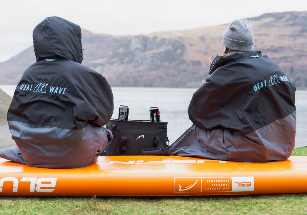 Two people in Heat Wave jackets sitting on an orange paddleboard by a lake, with drinks beside them.
