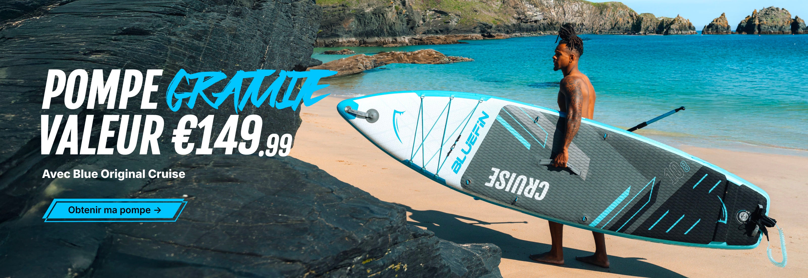 Man carrying a Bluefin inflatable paddleboard on a beach with rocky cliffs and blue water.
