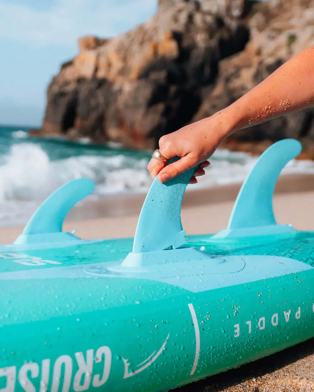 Hand adjusting the fin on a turquoise inflatable paddleboard on a sandy beach
