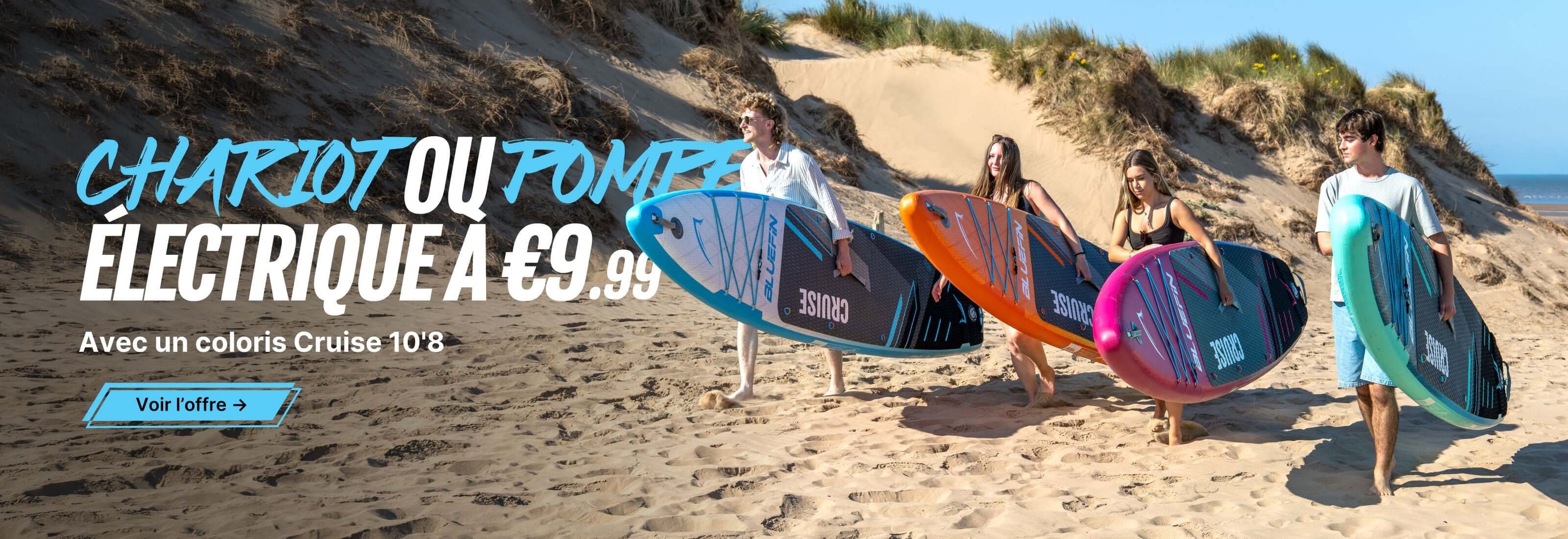 Four people carry colorful stand-up paddleboards on a sandy beach with dunes in the background.