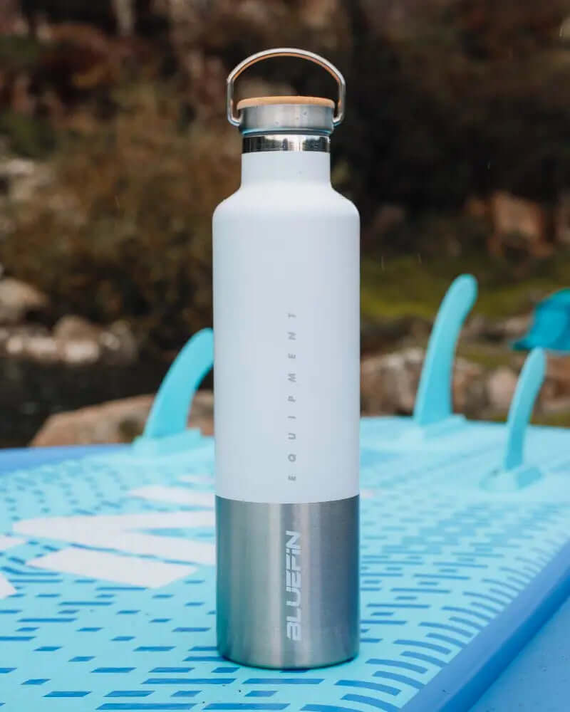 White insulated water bottle with stainless steel bottom, placed on a blue paddleboard.