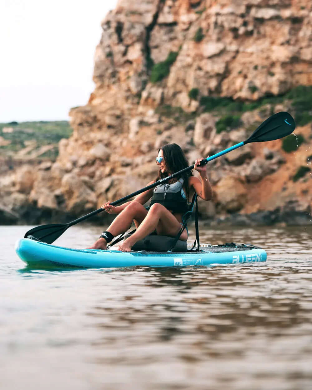 Woman paddling a blue inflatable paddleboard in calm water near rocky cliffs