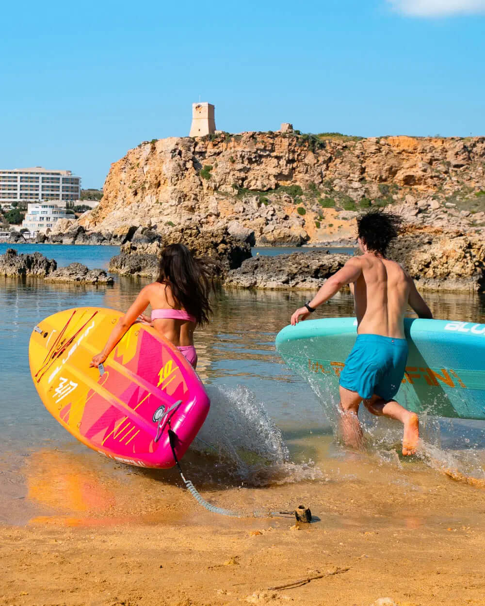 Two people running into the water with colorful paddleboards, rocky shore and tower in background.