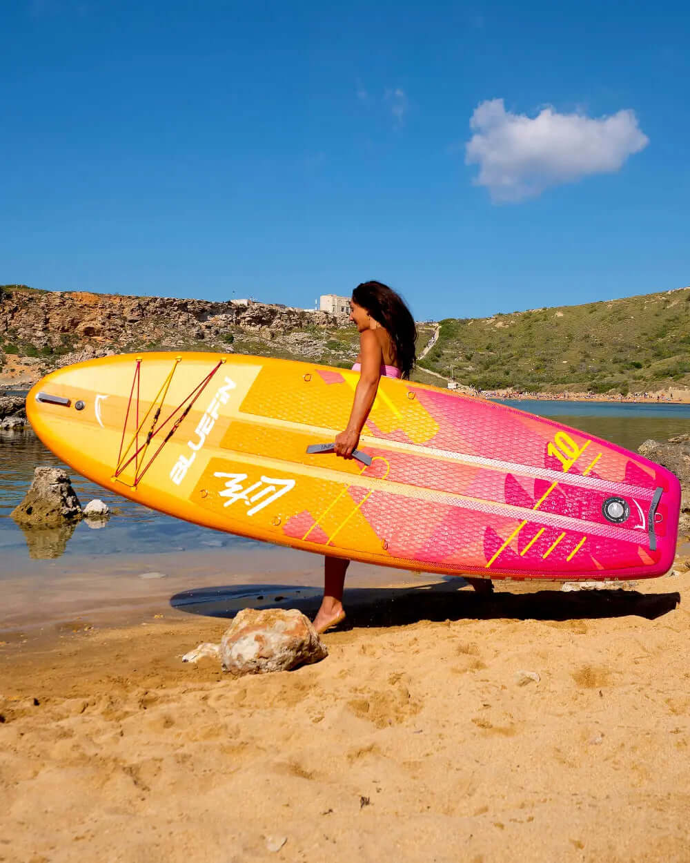 Person carrying a colorful Bluefin paddleboard on sandy beach near water
