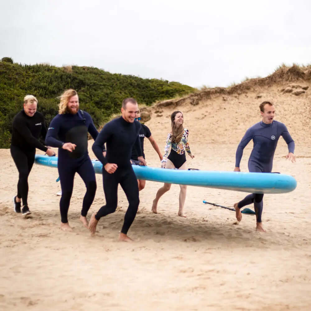 Groupe de six personnes en combinaisons de plongée courant sur le sable avec une grande planche de paddle bleue