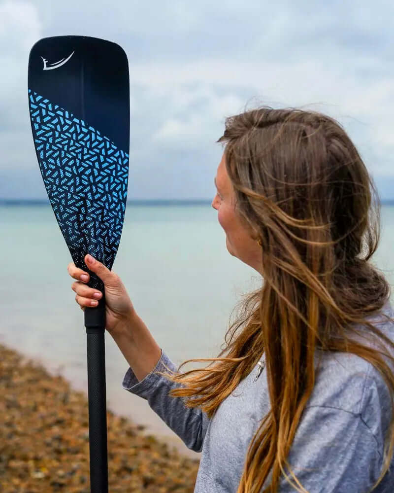 Person holding a black paddle with blue geometric design near a beach and calm water