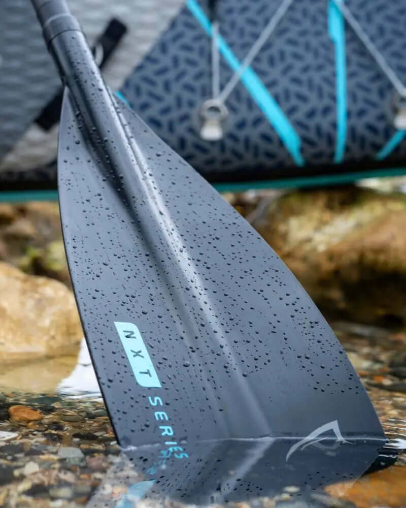 Close-up of a black paddle blade with water droplets, labeled NXT Series, beside a paddleboard.