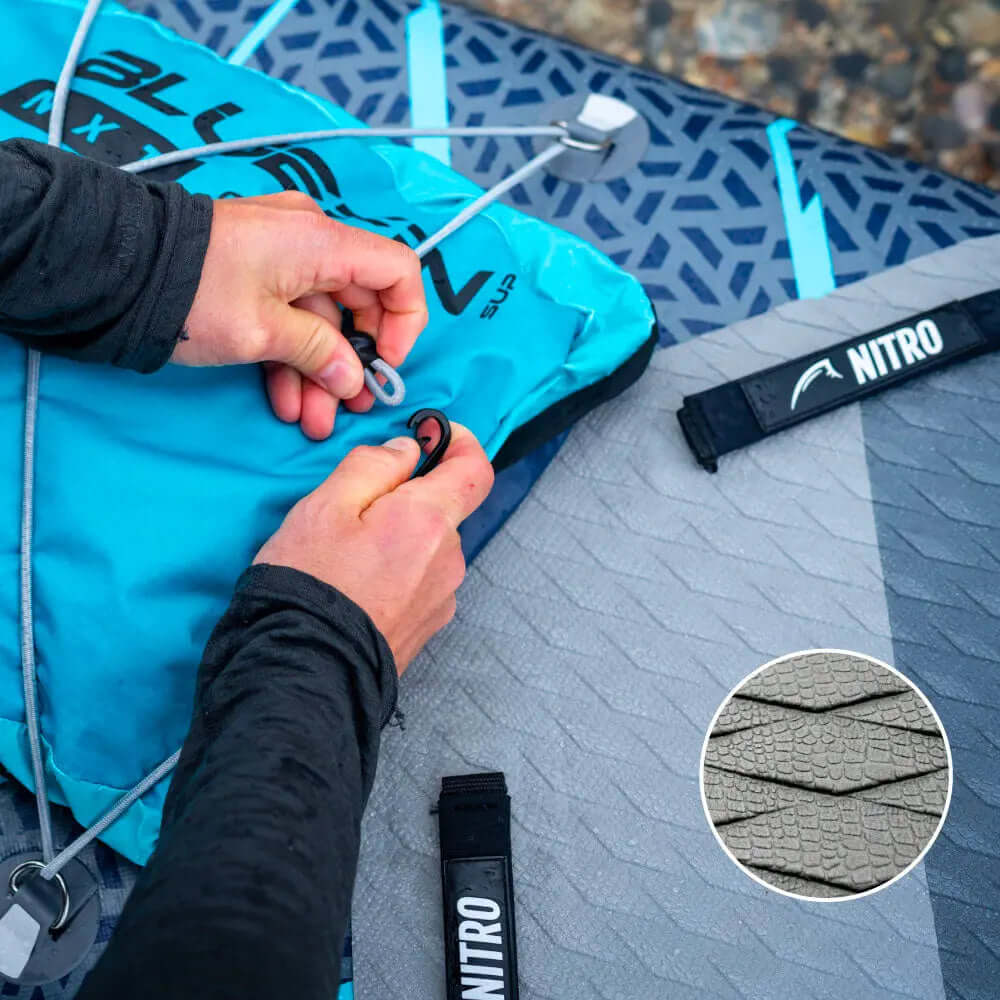 Mains sécurisant un sac de planche de paddle gonflable bleue avec un pad de pont texturé gris et une sangle NITRO.