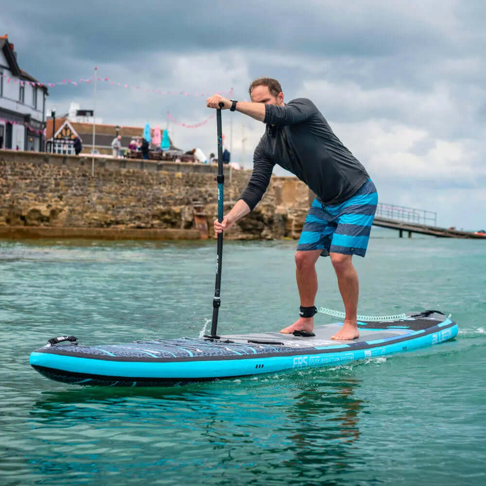 Homme pagayant sur une planche à pagaie gonflable bleue dans une eau calme, portant un t-shirt noir et un short rayé.