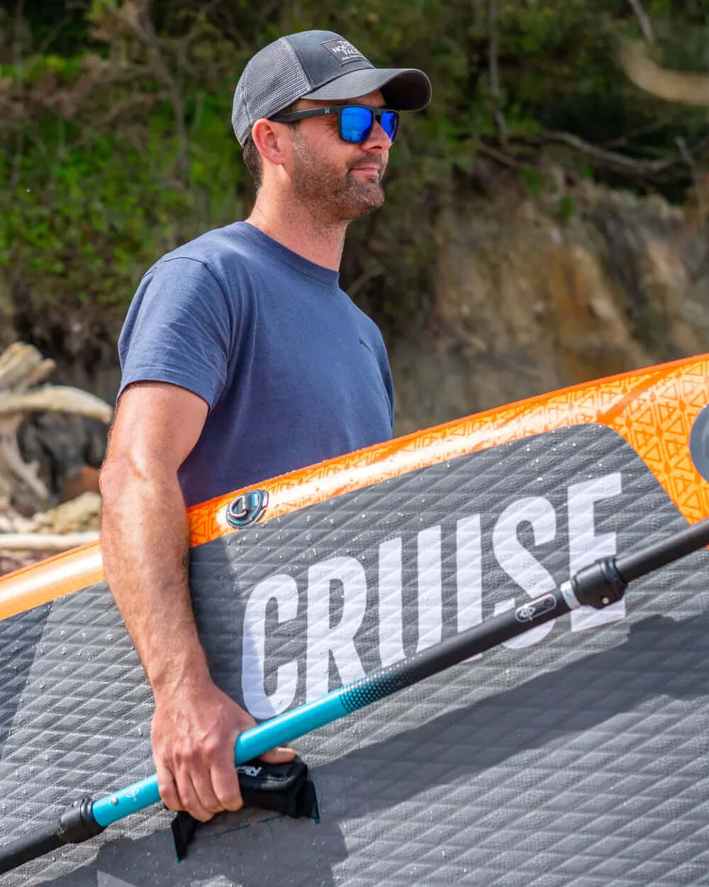 Man holding an inflatable paddleboard with orange edge and textured gray surface, wearing sunglasses and a cap.