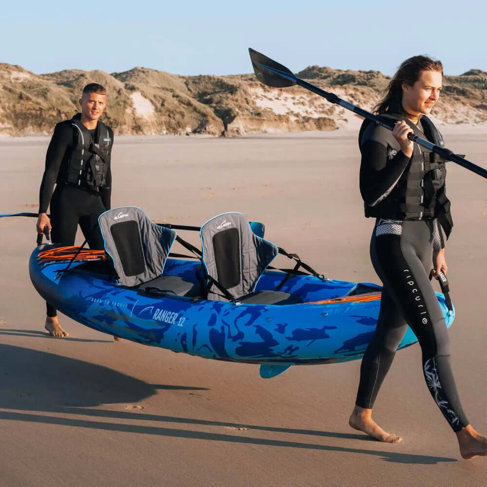 Two people walking on the beach with a blue inflatable kayak, one carrying a paddle.