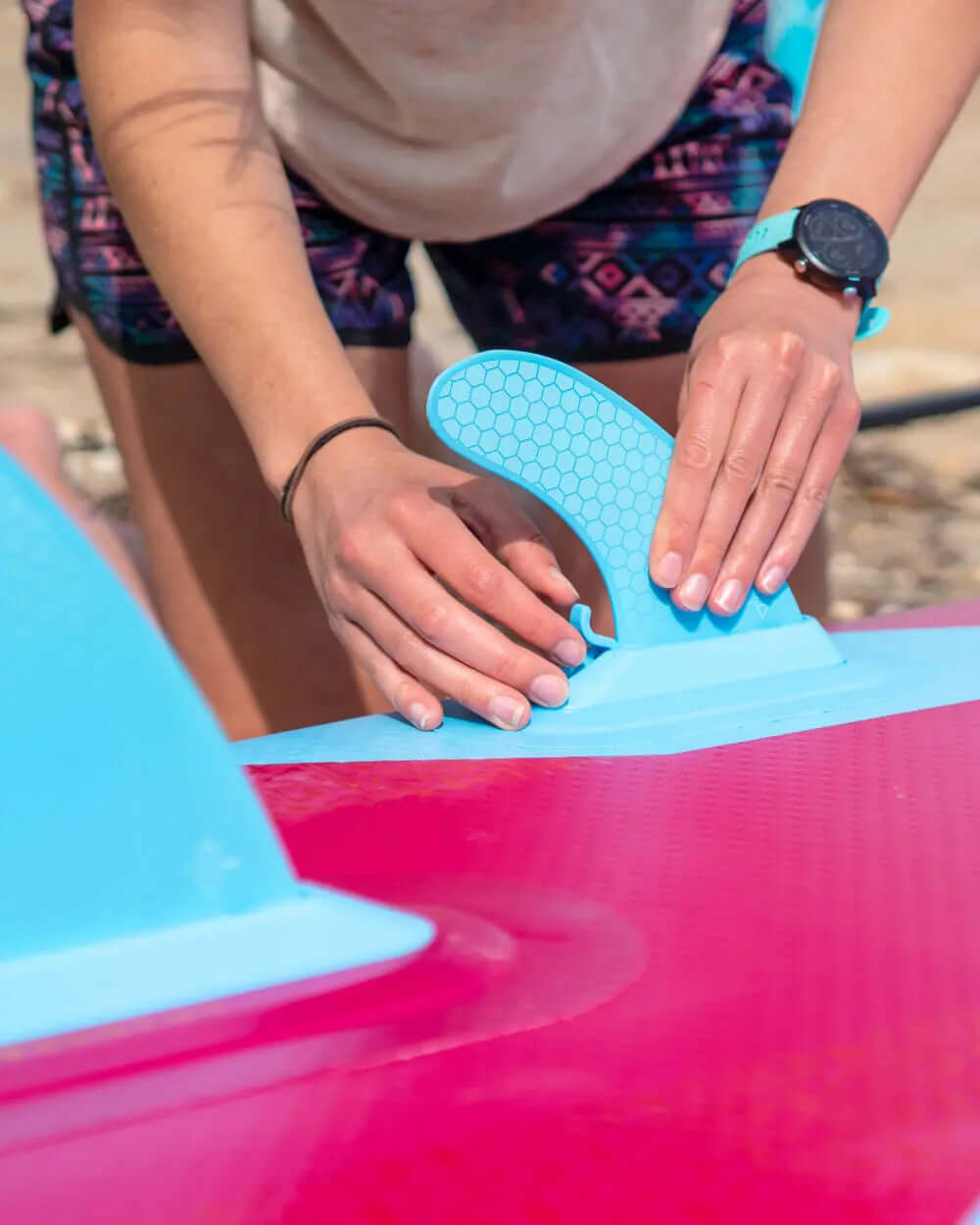Person attaching a blue fin to a pink inflatable paddleboard on a sunny beach