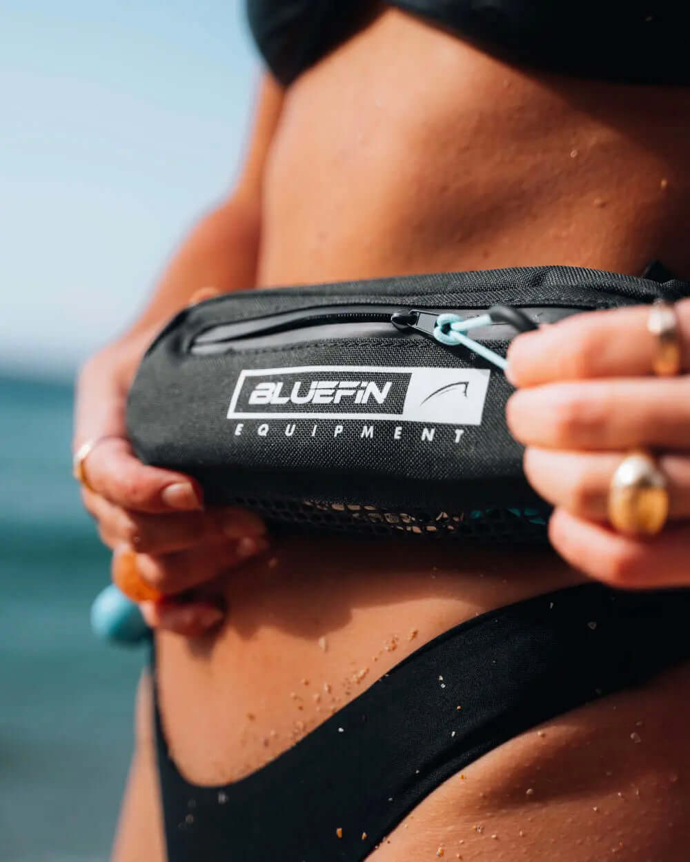 Person holding a black waterproof pouch with Bluefin Equipment logo, sandy beach in background