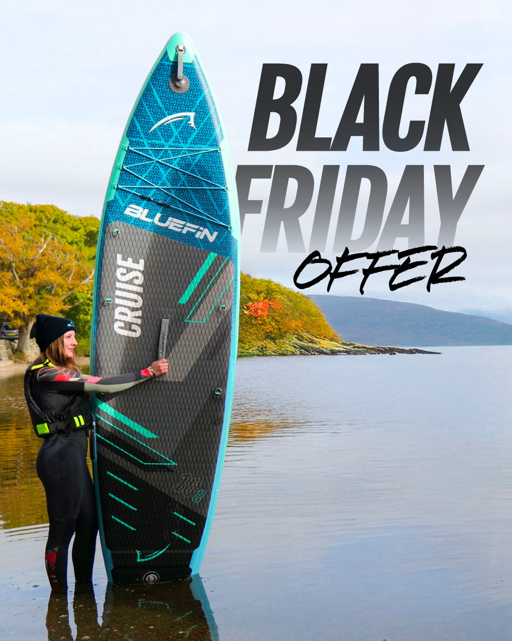Woman in wetsuit holding a Bluefin inflatable paddleboard in shallow water with autumn trees in background