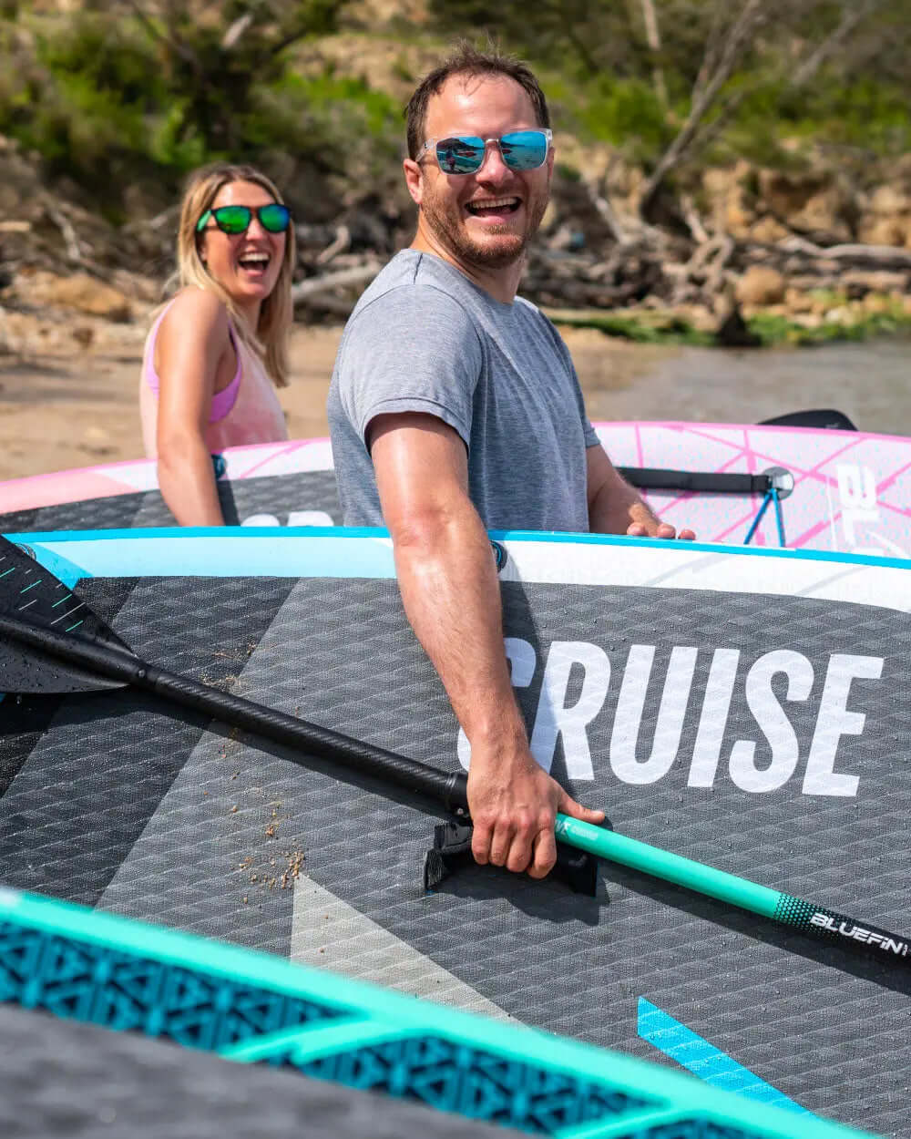 Two people smiling near inflatable paddleboards on a beach, one holding a paddle.
