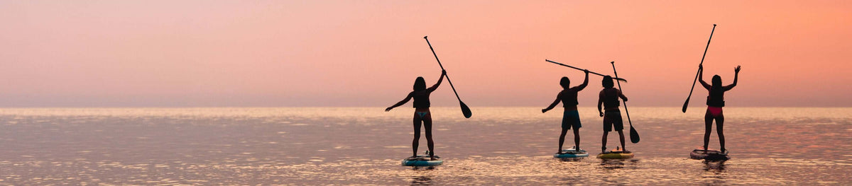 Quatre personnes faisant du paddle au coucher du soleil, en silhouette sur un ciel rose et une eau calme