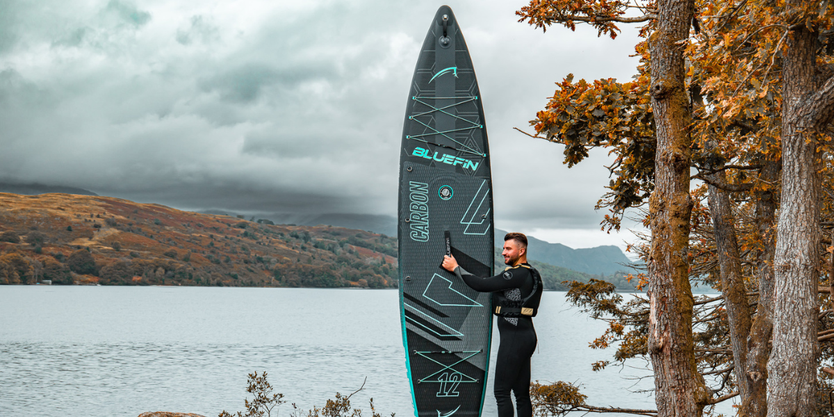 man holding a paddleboard infront of a lake 