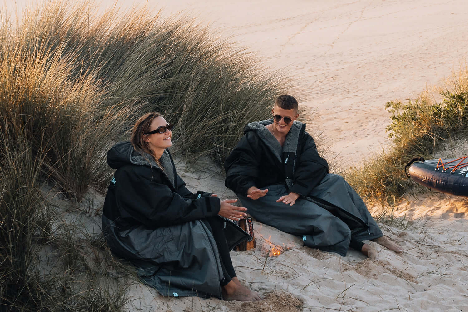 Two people sitting on the beach in oversized black jackets, laughing near a small fire.