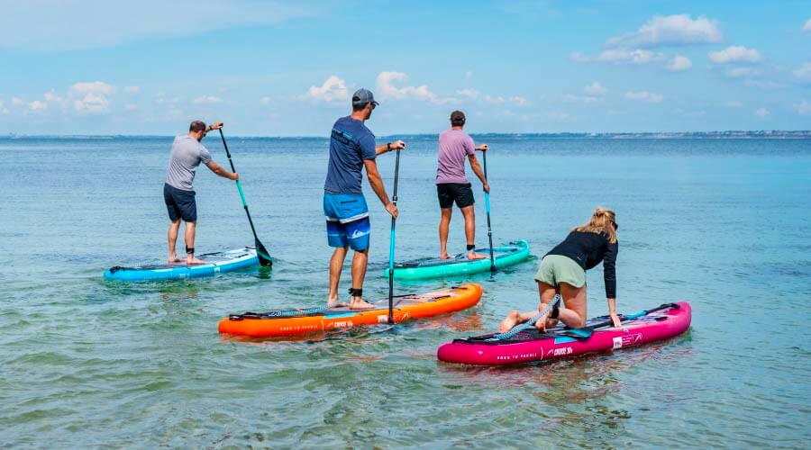 Four people paddleboarding on colorful inflatable boards in clear water under a blue sky.