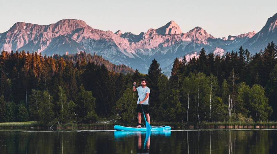 Man paddleboarding on a blue inflatable board in a lake with mountains in the background