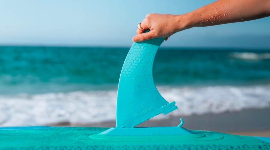 Hand holding a turquoise fin above a paddleboard on the beach with ocean waves in the background