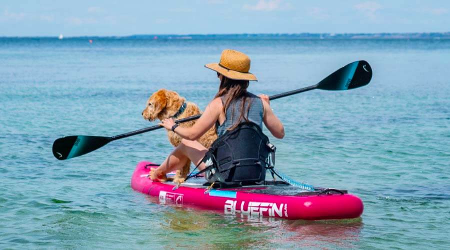 Woman paddling on a pink inflatable paddleboard with a dog, wearing a straw hat in clear water