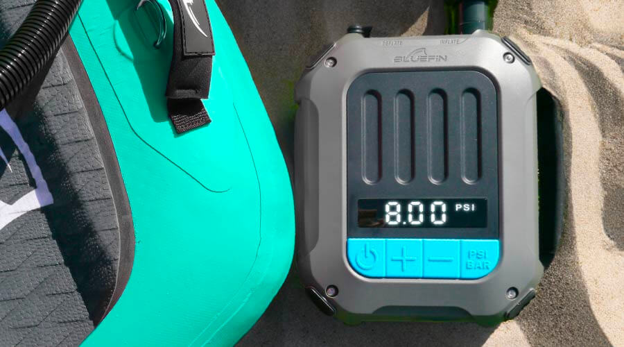Inflation pump displaying 8.00 PSI next to a teal inflatable paddleboard on sand