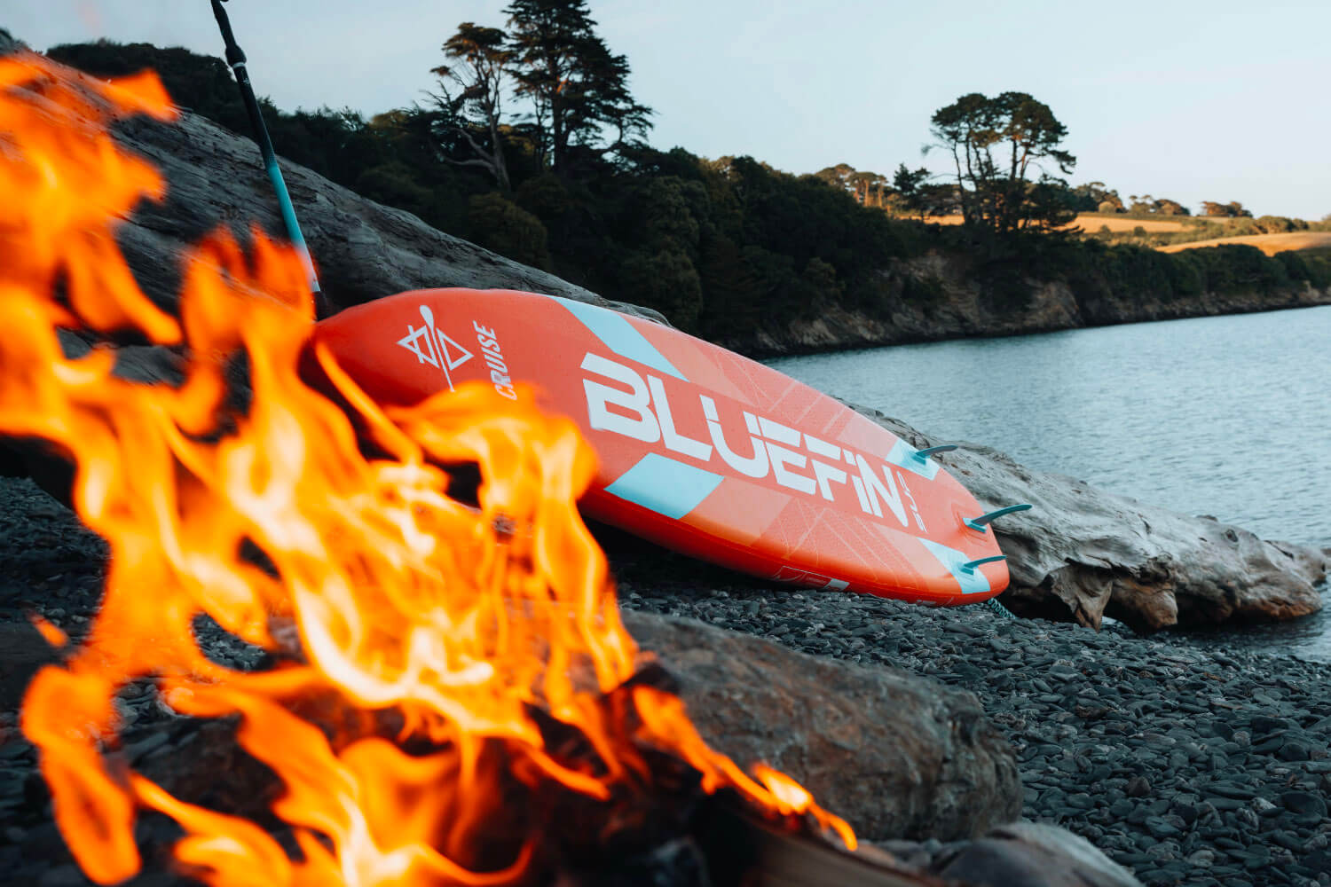 Inflatable paddleboard with orange and blue design resting on rocky shore near a fire and water.