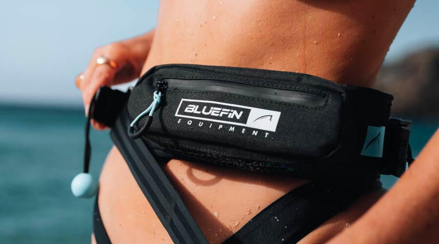 Close-up of a person wearing a Bluefin waist pack by the water, showing logo and adjustable strap.